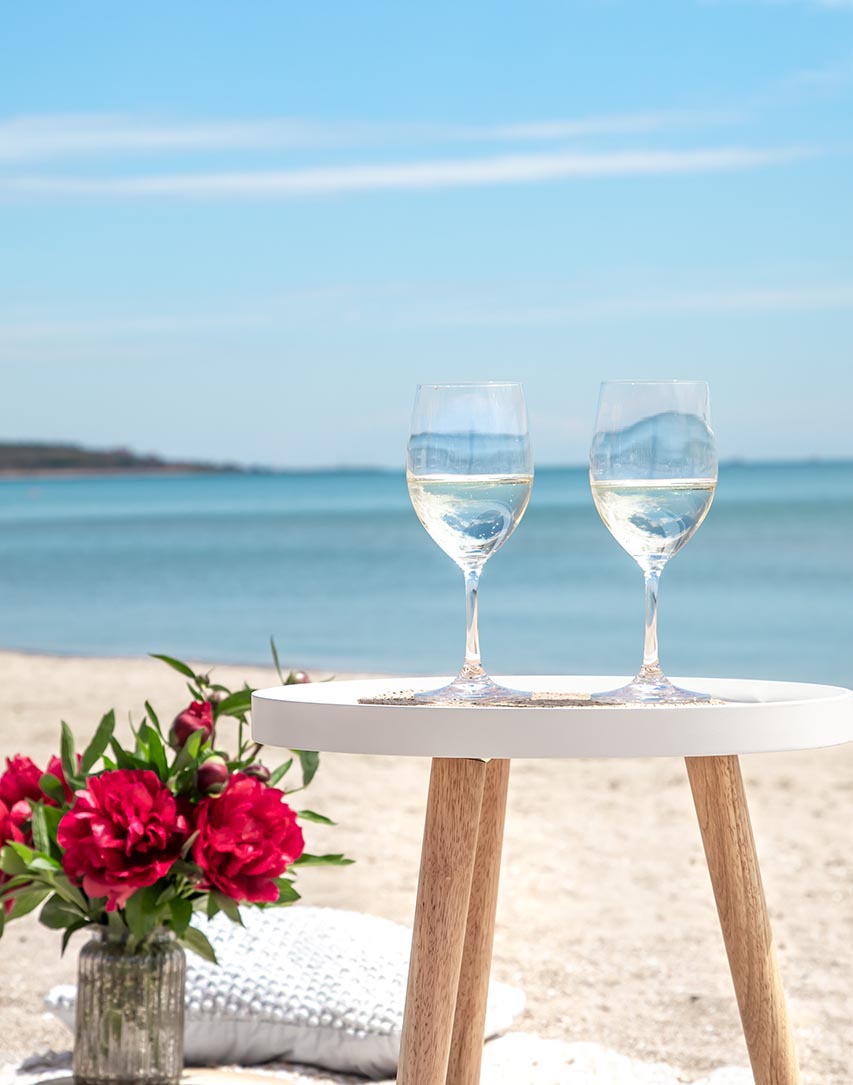 Two wine glasses on a small table by the beach with bright red flowers and ocean views, creating a romantic seaside setting.
